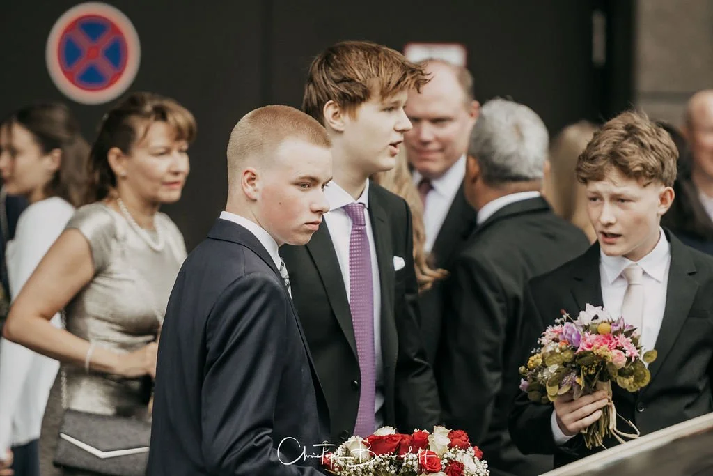 Jugendliche in formeller Kleidung bei einer Feier, einige mit Blumensträußen, im Hintergrund weitere Menschen.