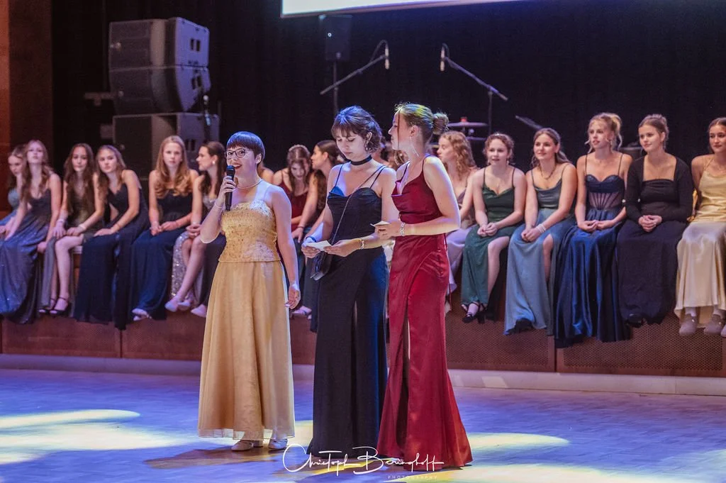 Junge Frauen in eleganten Abendkleidern sitzen auf einer Empore bei einer Veranstaltung, während drei Frauen vorne sprechen, eine hält ein Mikrofon. Im Hintergrund sind Mikrofongeräte sichtbar.