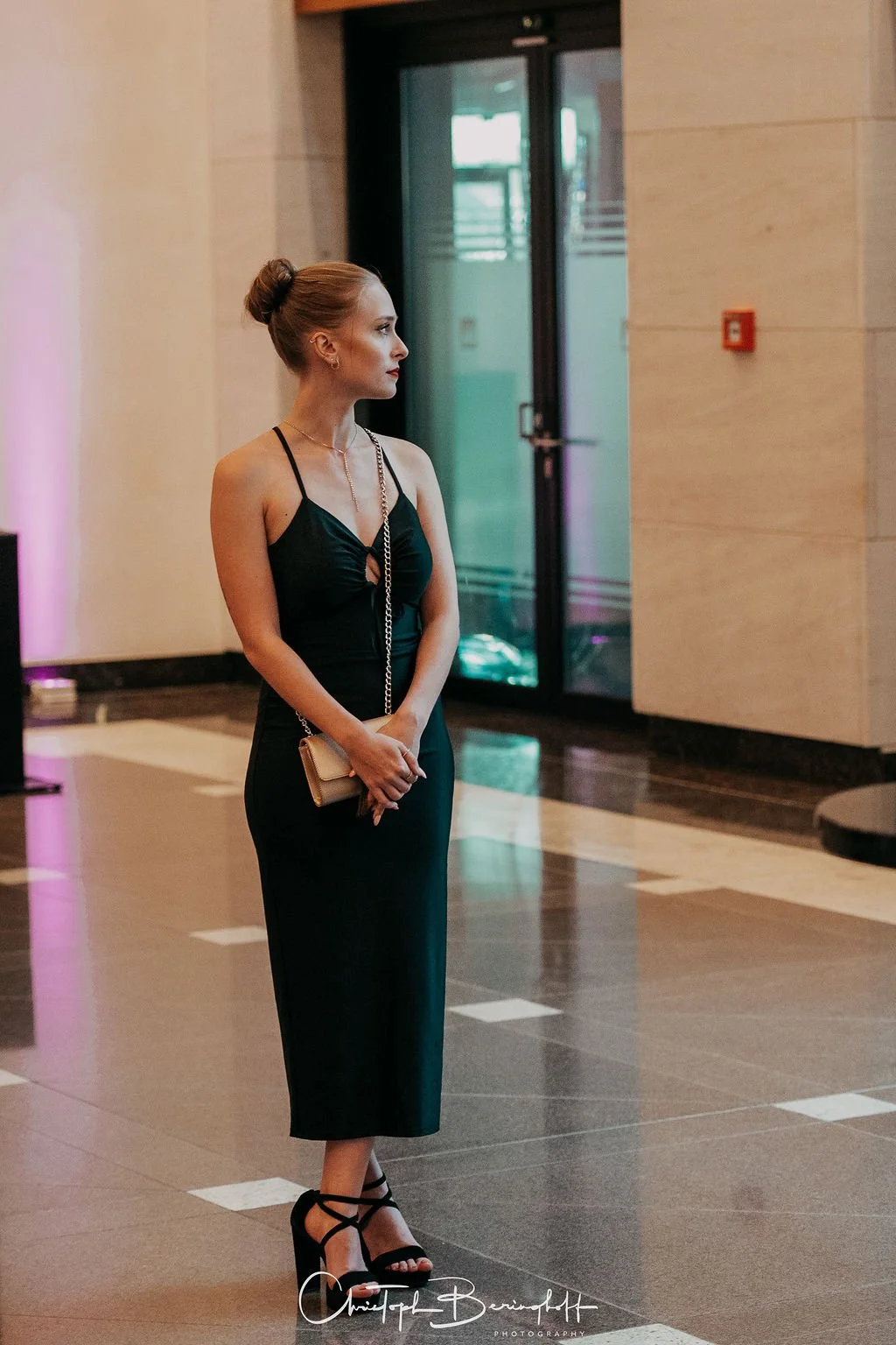 Eine elegante Frau trägt ein schwarzes Kleid mit dünnen Trägern, schwarze High Heels und eine kleine silberne Handtasche, steht in einer modernen Lobby.