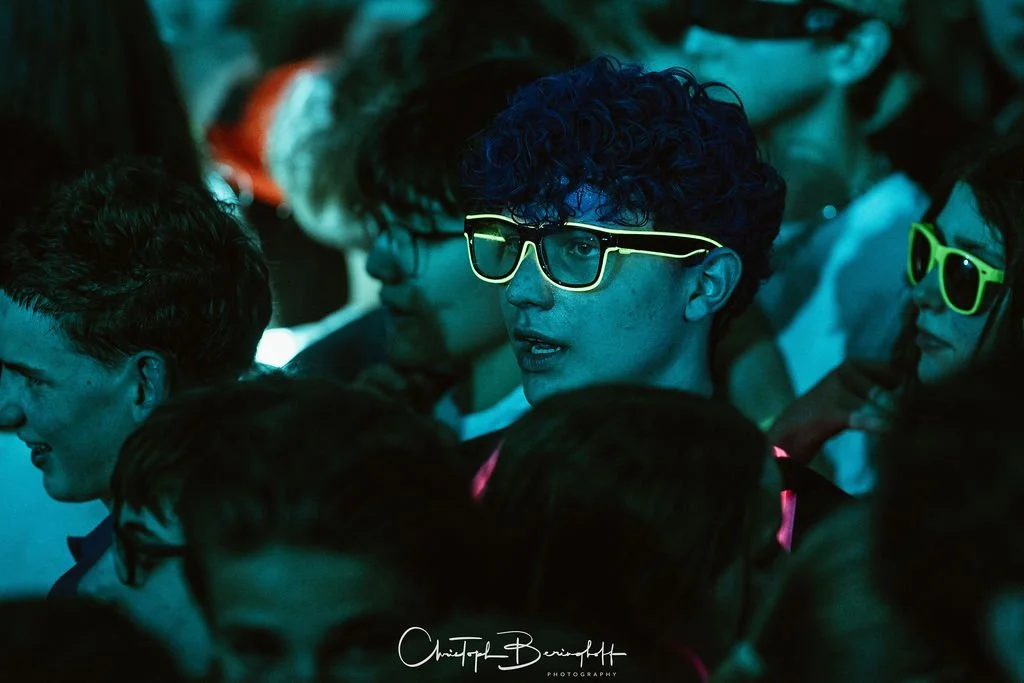 People at an event, some wearing bright yellow and green sunglasses, against a dark background with blue lighting.