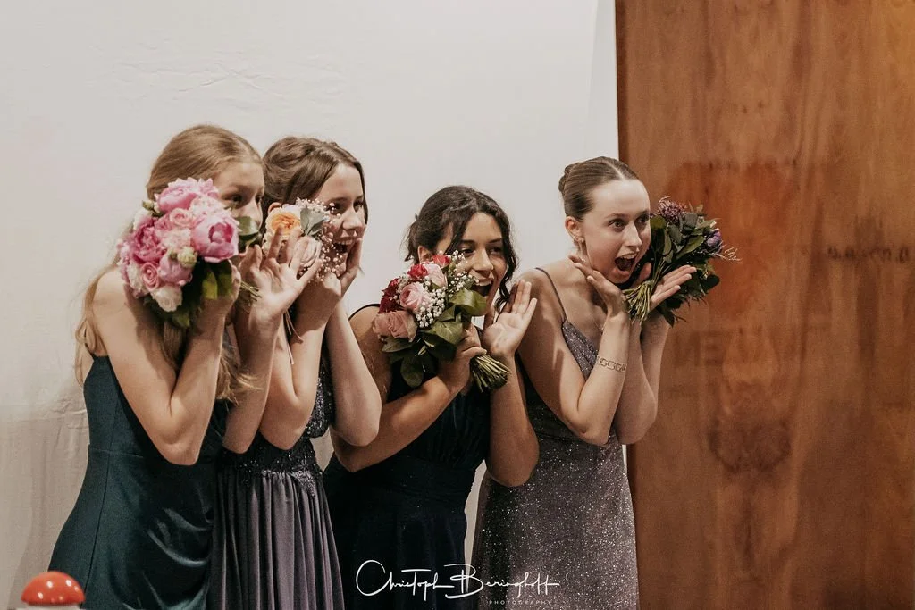 Vier junge Frauen in eleganten Kleidern, die im Freien Blumen halten und überraschte, freudige Gesichter zeigen.