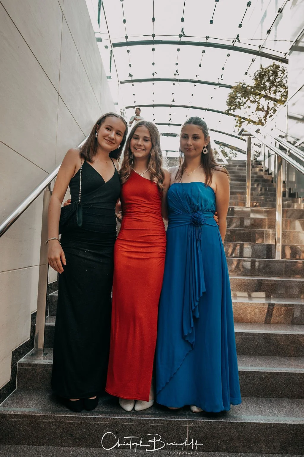 Drei junge Frauen in elegante Abendkleider stehen auf einer Treppe in einem modernen Gebäude, lächelnd und nebeneinander posierend.