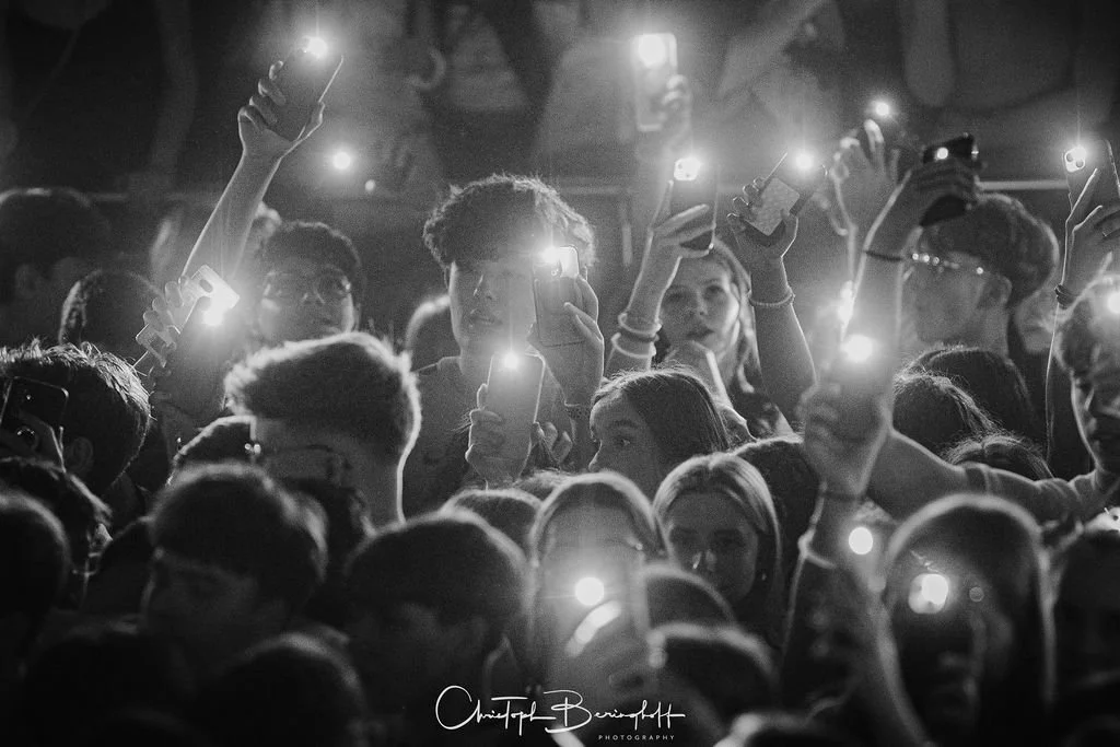 A crowd at a concert, many holding up their cell phones, black-and-white photo.