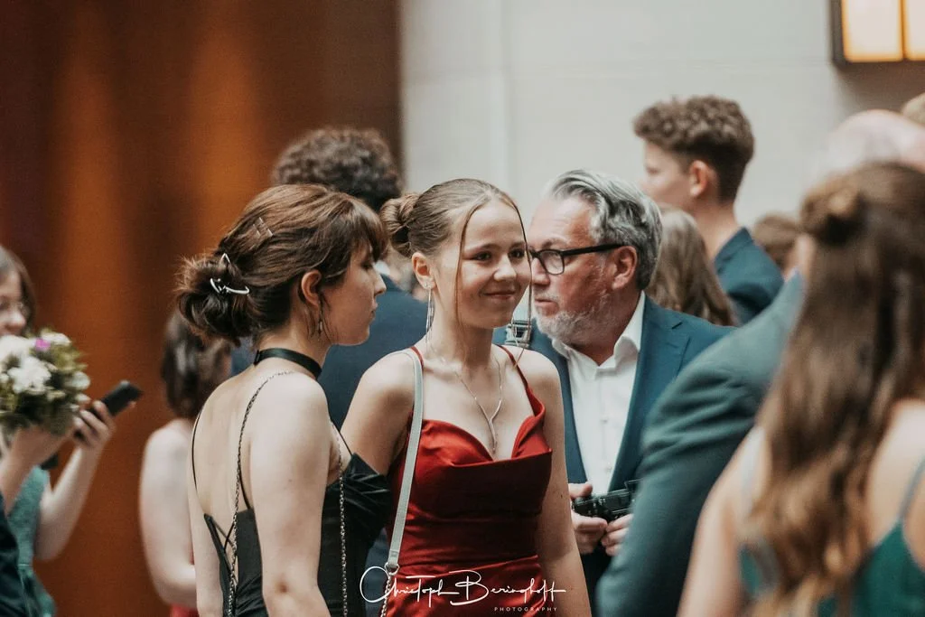Junge Frau in rotem Kleid bei einer Veranstaltung, umgeben von anderen, im Gespräch mit einem Mann mit Brille.