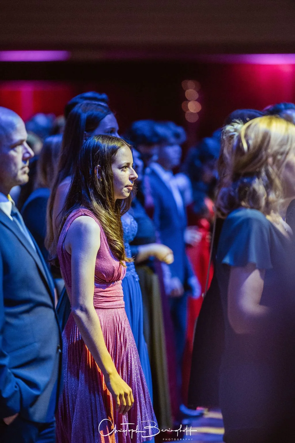 Menschen bei einer formellen Veranstaltung, die in einer dunklen Umgebung mit buntem Licht sitzen oder stehen, einige tragen elegante Kleidung.
