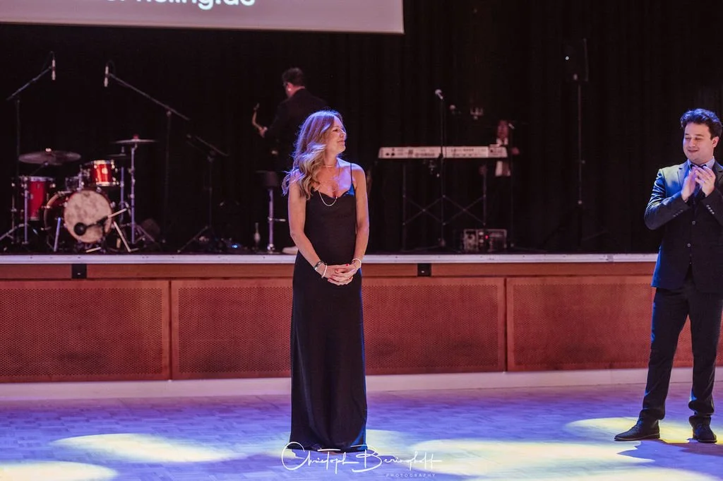 Eine Frau in einem schwarzen Abendkleid steht auf einer Bühne, neben einem Mann in einem dunklen Anzug, bei einer Veranstaltung mit einem Musiksetup im Hintergrund.
