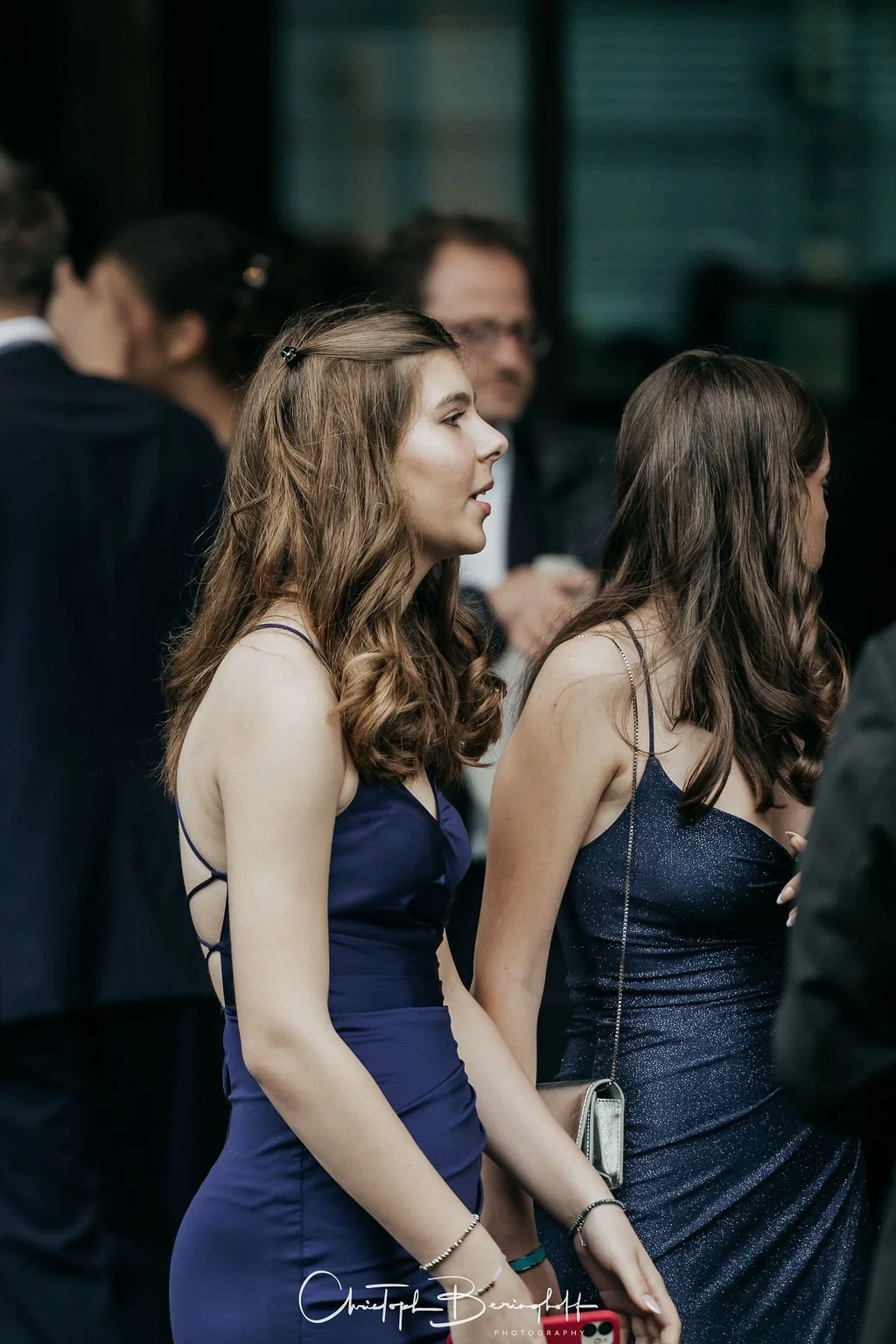 Zwei junge Frauen in eleganten dunklen Kleidern bei einer formellen Veranstaltung, im Hintergrund mehrere Personen.