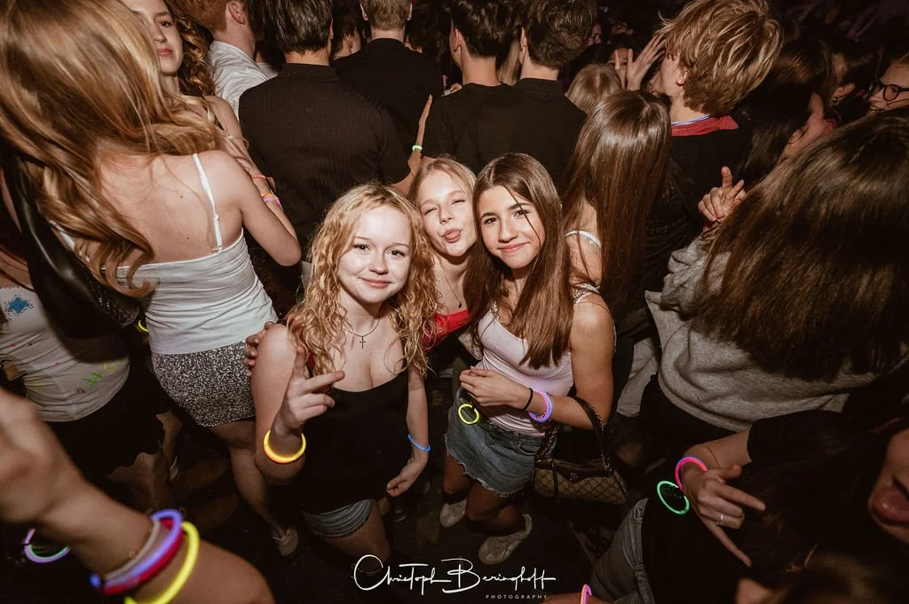 Three young women are dancing and posing in a crowd at a party or event, wearing summer outfits and neon jewelry.
