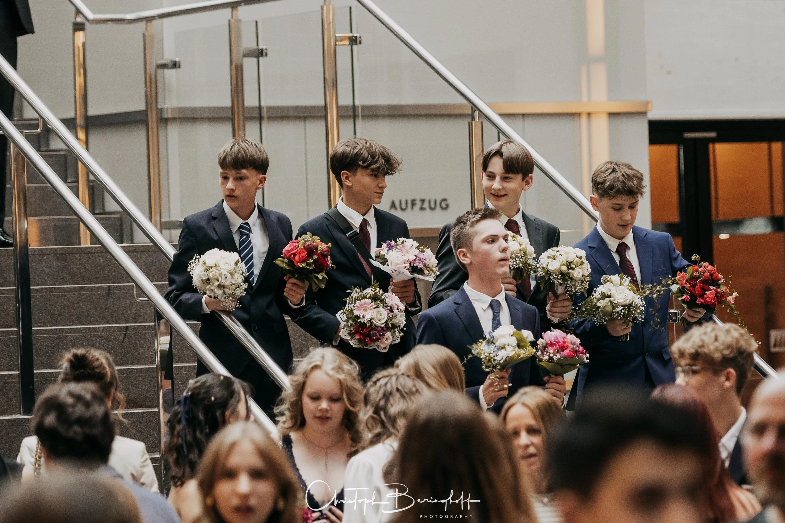 Junge Männer in Anzügen mit Blumen in den Händen, die beim Eintreten in einen Saal für eine Zeremonie oder Feier auf der Treppe stehen.