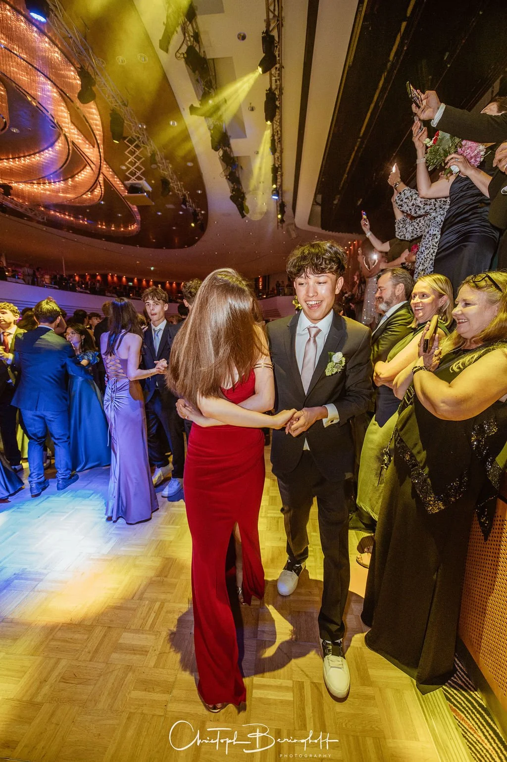 Menschen tanzen auf einer festlichen Veranstaltung, die im eleganten Saal mit bunten Lichtern und Damen in eleganten Kleidern stattfindet.