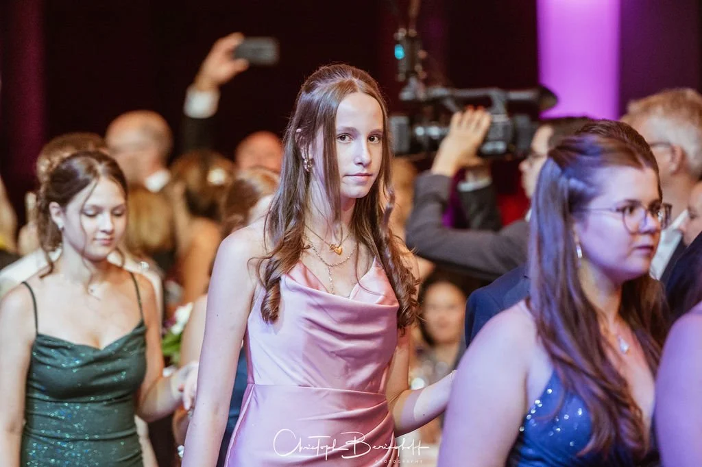 Junge Frau in rosa Abendkleid bei einer Gala oder Feier, umgeben von anderen Gästen.