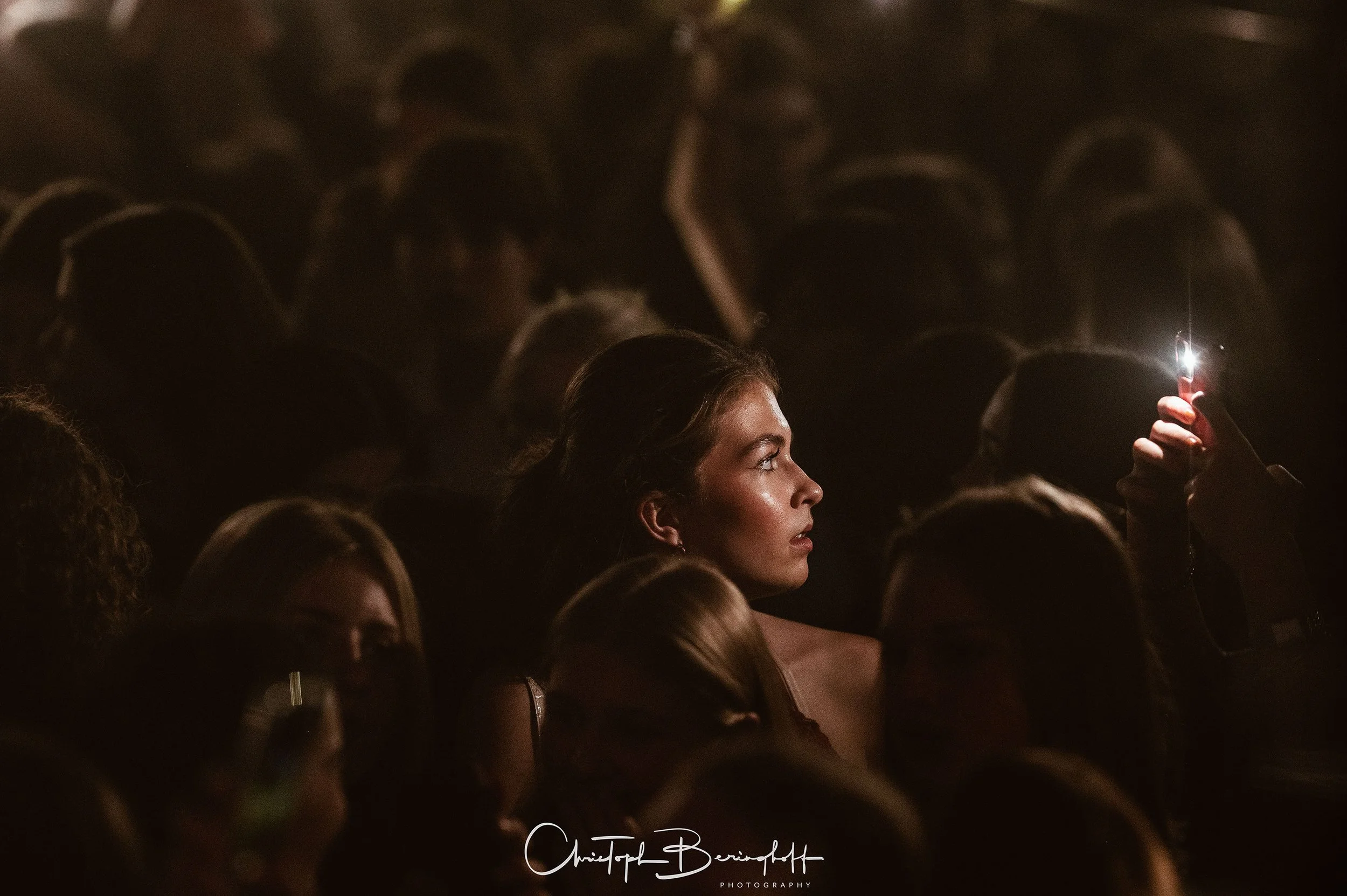 Eine junge Frau mit schimmernder Haut sitzt in einer dunklen Umgebung, umgeben von anderen Menschen, einige tragen Masken. Sie hält ihr Smartphone hoch und leuchtet es auf, während sie aufmerksam auf etwas richtet.