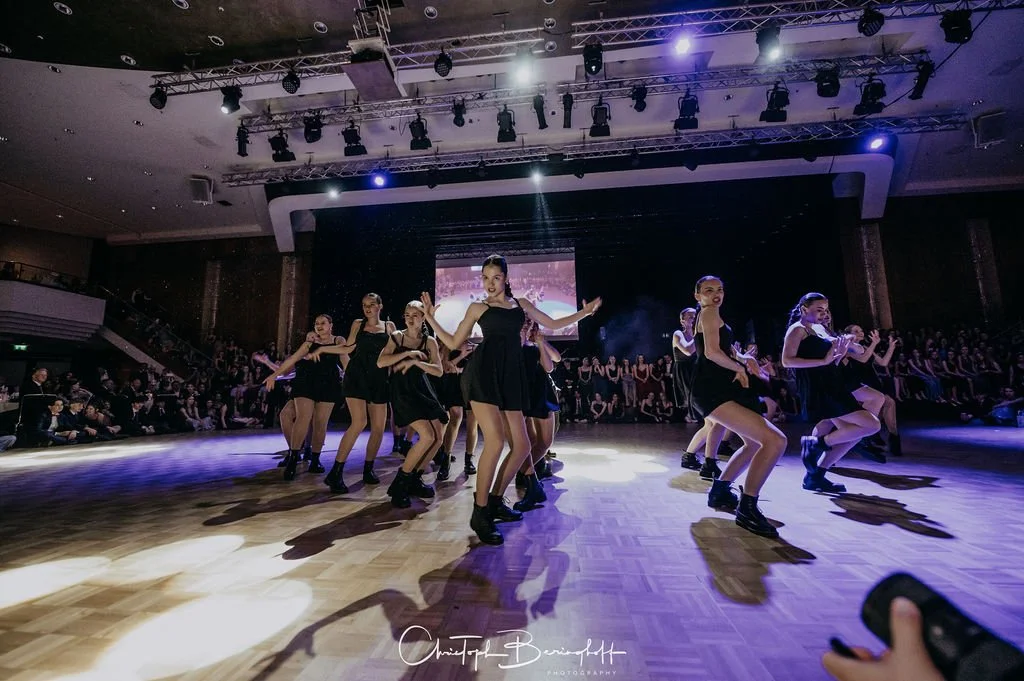 Tänzer auf einer Bühne in Schwarz tanzend während eines Performances vor Publikum, mit Bühnenbeleuchtung und Zuschauern im Hintergrund.