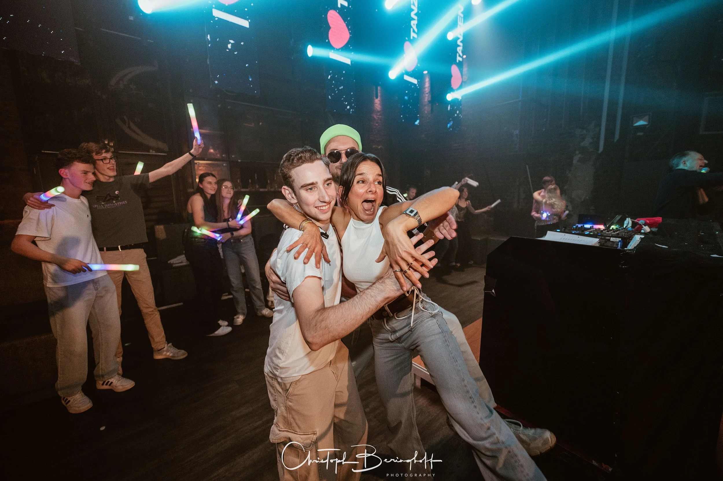 Menschen feiern auf einer Party im Dunkeln, einige tanzen mit leuchtenden Lichtstäben, während ein DJ am DJ-Pult Musik auflegt. Die Stimmung ist lebhaft und fröhlich, mit buntem Licht und Begeisterung bei den Gästen.