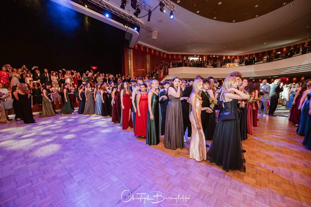 Eine Gruppe von jungen Menschen, die in eleganten Abendkleidern und Anzügen bei einer formellen Veranstaltung auf einem Holzboden im Saal stehen, mit vielen Zuschauern im Hintergrund.