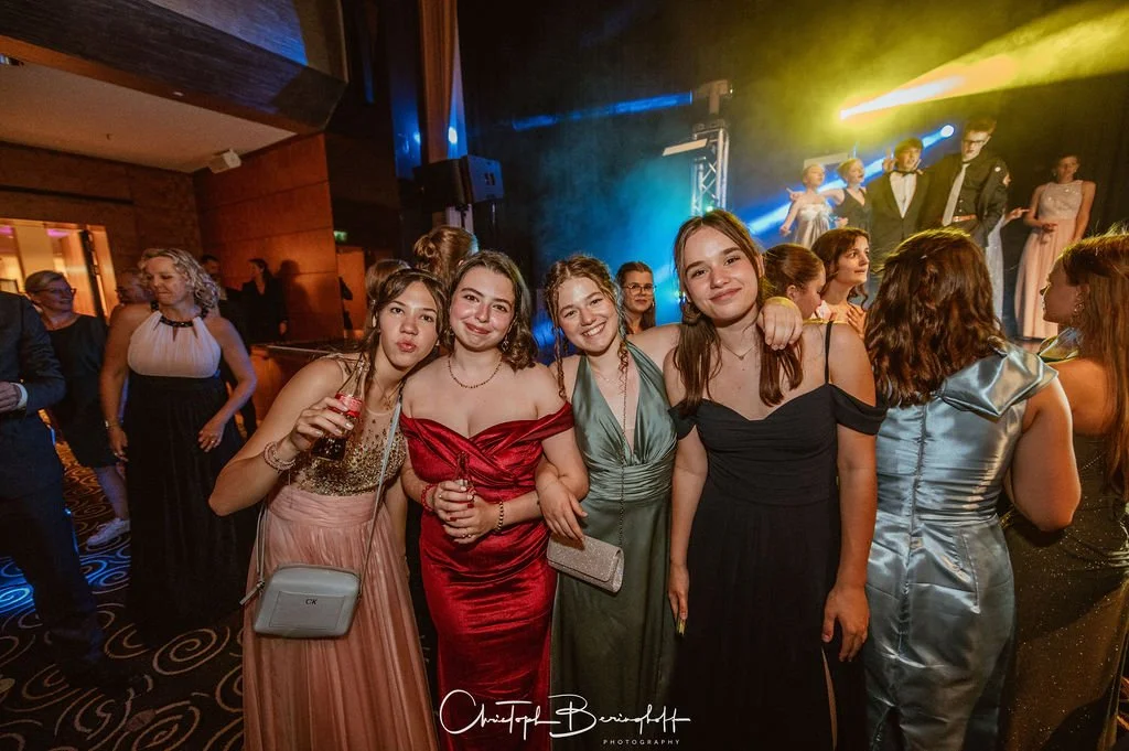 Gruppe junger Frauen in eleganten Kleidern feiern auf einer Party, mit einem DJ-Setup im Hintergrund und bunten Lichtern.