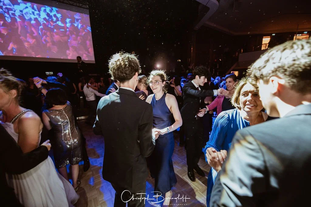 Menschen tanzen und feiern auf einer Gala mit dunklem Raum, großer Leinwand im Hintergrund und Disco-Atmosphäre.