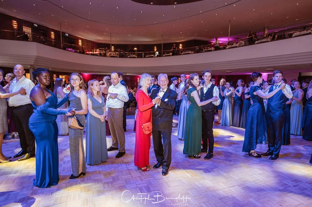Menschen tanzen in einem großen Saal, viele sind in eleganter Abendkleidung. Die Atmosphäre ist festlich mit bunten Lichtern, der Saal ist zweistöckig mit einer oberen Balustrade.