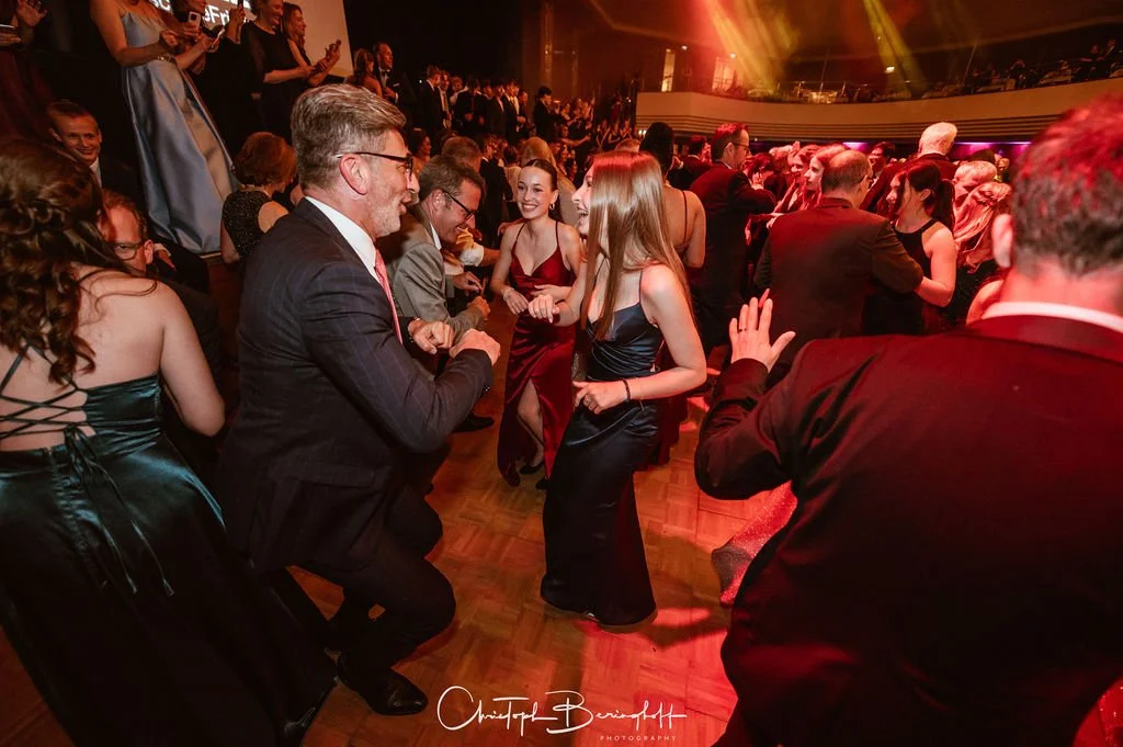 Tanzende Menschen bei einer festlichen Veranstaltung, überwiegend in eleganter Kleidung, auf einer Tanzfläche mit bunten Lichtern im Hintergrund.