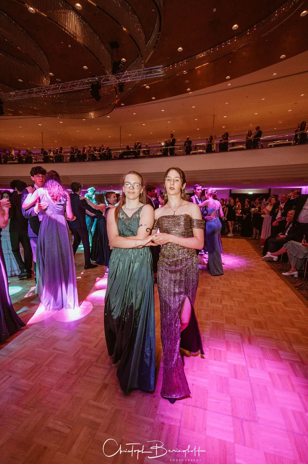 Zwei junge Frauen tanzen auf einer eleganten Party, umgeben von weiteren Gästen in Abendkleidern, in einem großen festlichen Saal mit Holzparkett und bunten Lichteffekten.