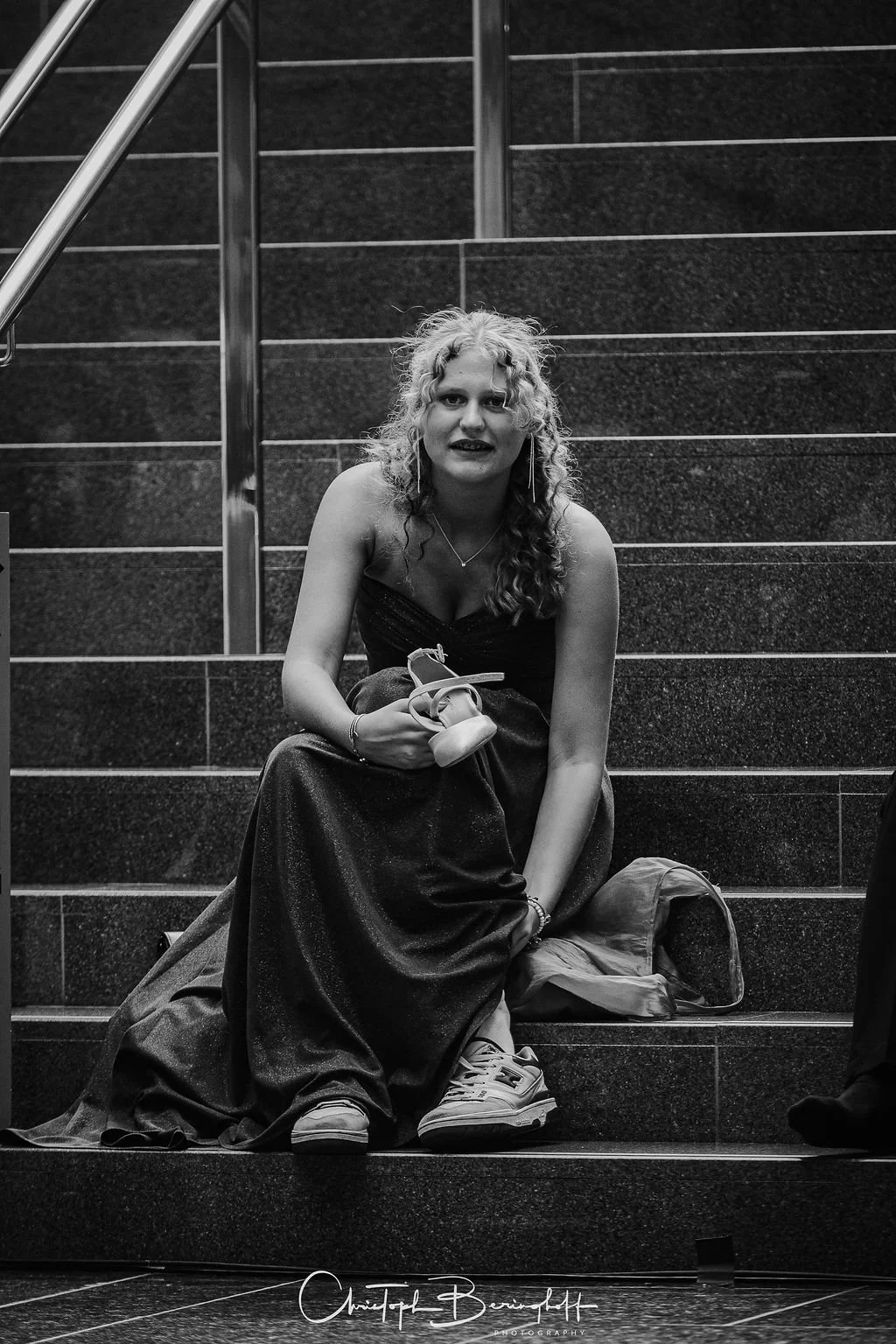 Junge Frau mit lockigem Haar, schwarze Abendkleidung, sitzt auf Treppe in öffentlichen Raum, hält Schuhe in der Hand, trägt Sneakers, neben Tasche und Jacke.