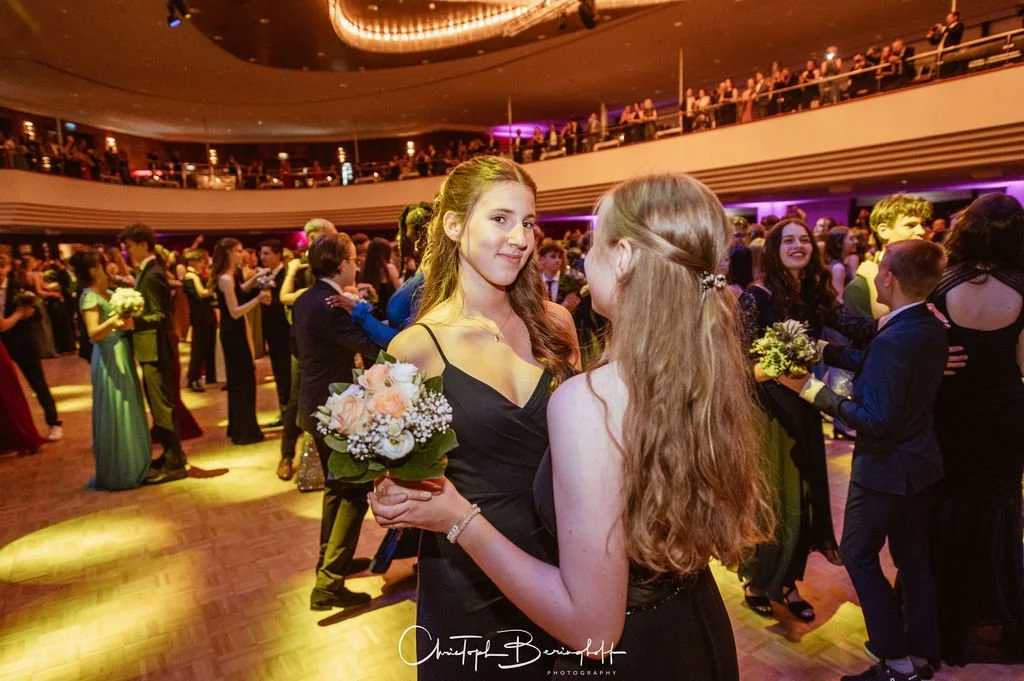 Junge Frauen tanzen bei einer formellen Veranstaltung mit vielen anderen Gästen auf der Tanzfläche.