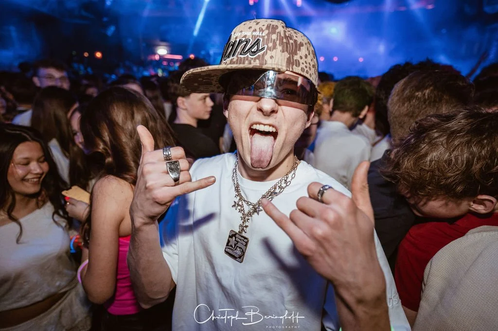 A young man wearing a camouflage cap, sunglasses, and necklaces makes a hand gesture, opens his mouth, and sticks out his tongue at a party.