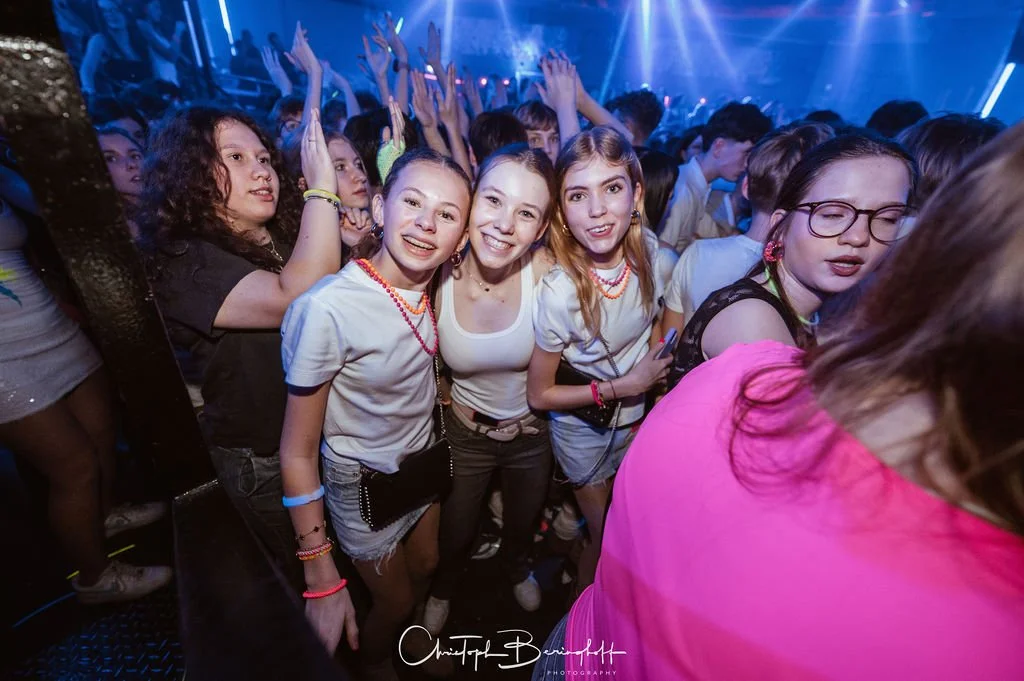 People are celebrating at a party with colorful lights; some are laughing, others are dancing.