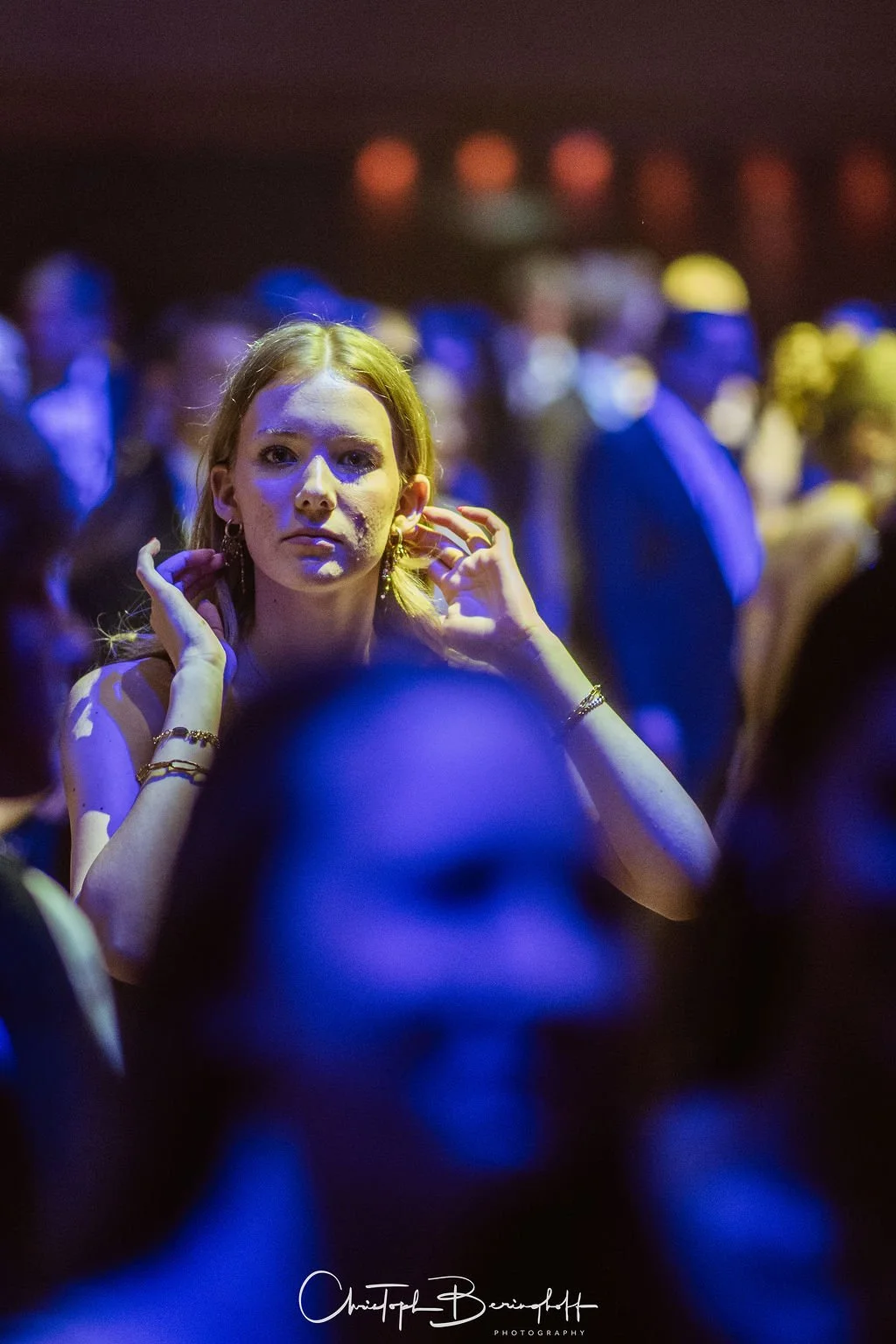 Junge Frau mit rotem Haar und Eheringen im Ohr sitzt unter buntem, gedämpftem Licht auf einer Party oder im Club, umgeben von anderen Menschen.