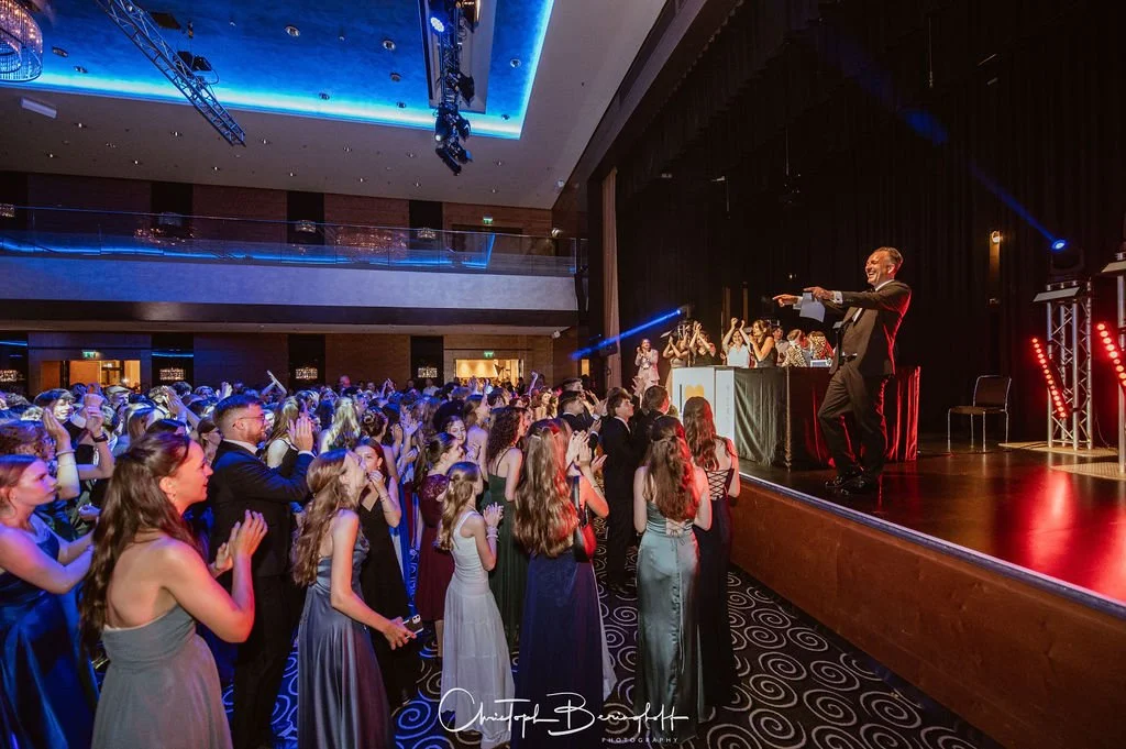 Menschen auf einer Tanzfläche bei einer Feier im eleganten Saal, im Hintergrund ein Mann auf einer Bühne mit einem Mikrofon, farbiges Licht und Vorhänge, festliche Atmosphäre.