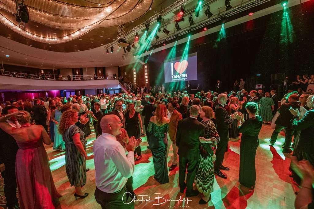 Tanzveranstaltung in einem großen Saal mit tanzenden Menschen, buntem Licht und einer großen Leinwand, auf der steht "I ♥ TANZEN".