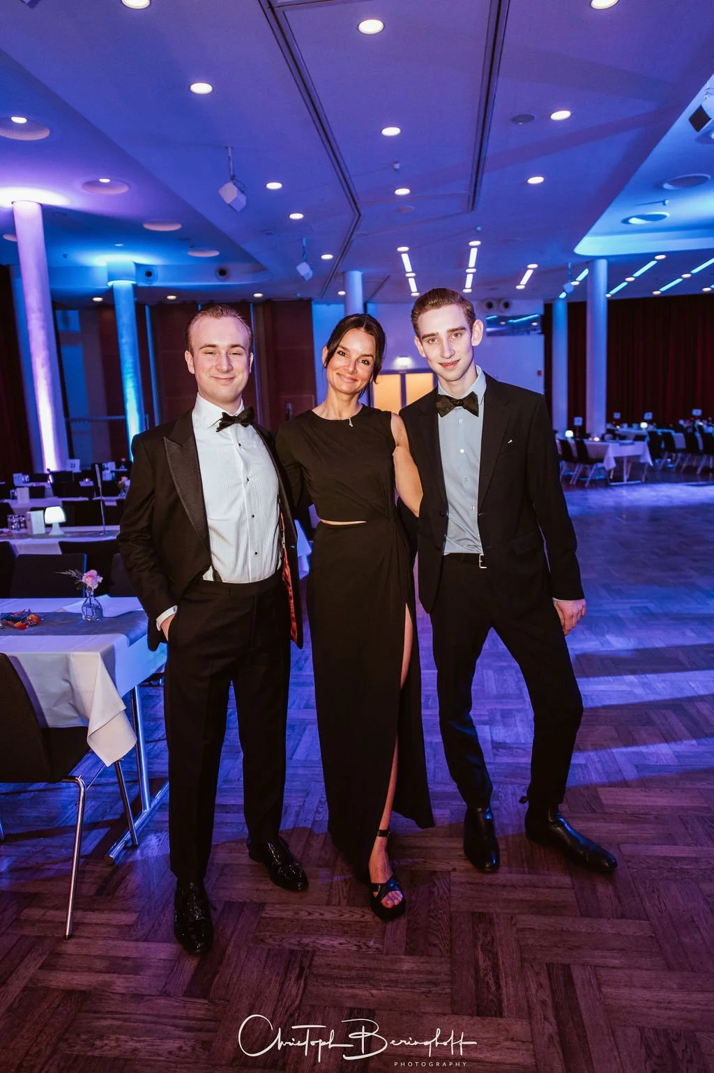 Drei Menschen in formeller Abendkleidung auf einer Tanzfläche bei einer eleganten Veranstaltung. Zwei Männer in schwarzen Anzügen und eine Frau in einem schwarzen Kleid mit hohem Schlitz stehen im Mittelpunkt.