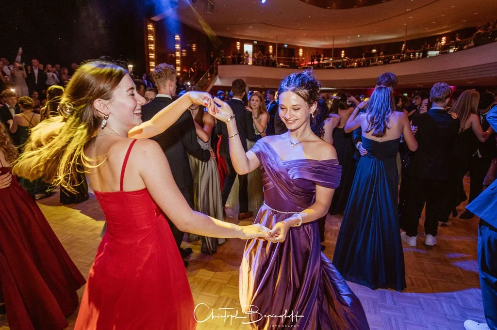 Zwei junge Frauen tanzen auf einer Party und lachen. Sie tragen elegante Abendkleider, eine in Rot und die andere in Lila, während sich im Hintergrund weitere Gäste in formeller Kleidung auf der Tanzfläche befinden.