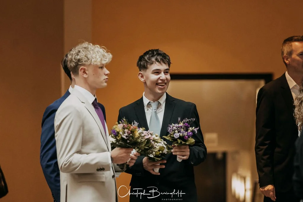 Zwei junge Männer in Anzügen halten Blumen und lachen in einem festlichen Raum.