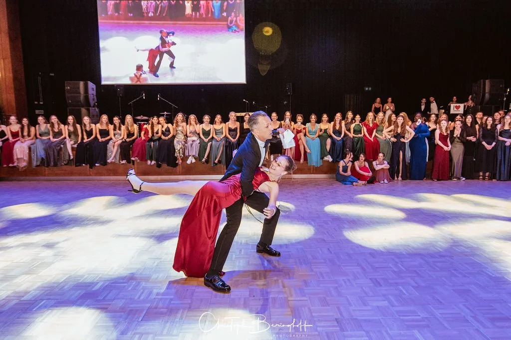 Tanzpaar bei einem Ballsaal-Tanz vor einem Publikum auf der Bühne mit großem Bildschirm im Hintergrund.