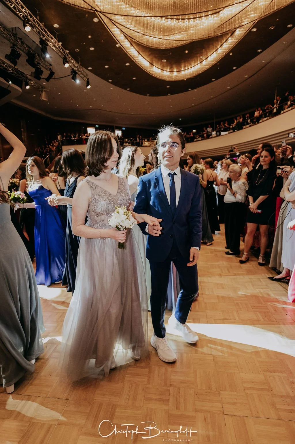 Menschen bei einer Hochzeitsfeier, im Vordergrund zwei junge Frauen, eine in einem langen grauen Kleid mit Blumenstrauß und die andere in einem blauen Anzug, Hand in Hand, auf einem Tanzparkett, im Hintergrund weitere Gäste in eleganter Kleidung, im 