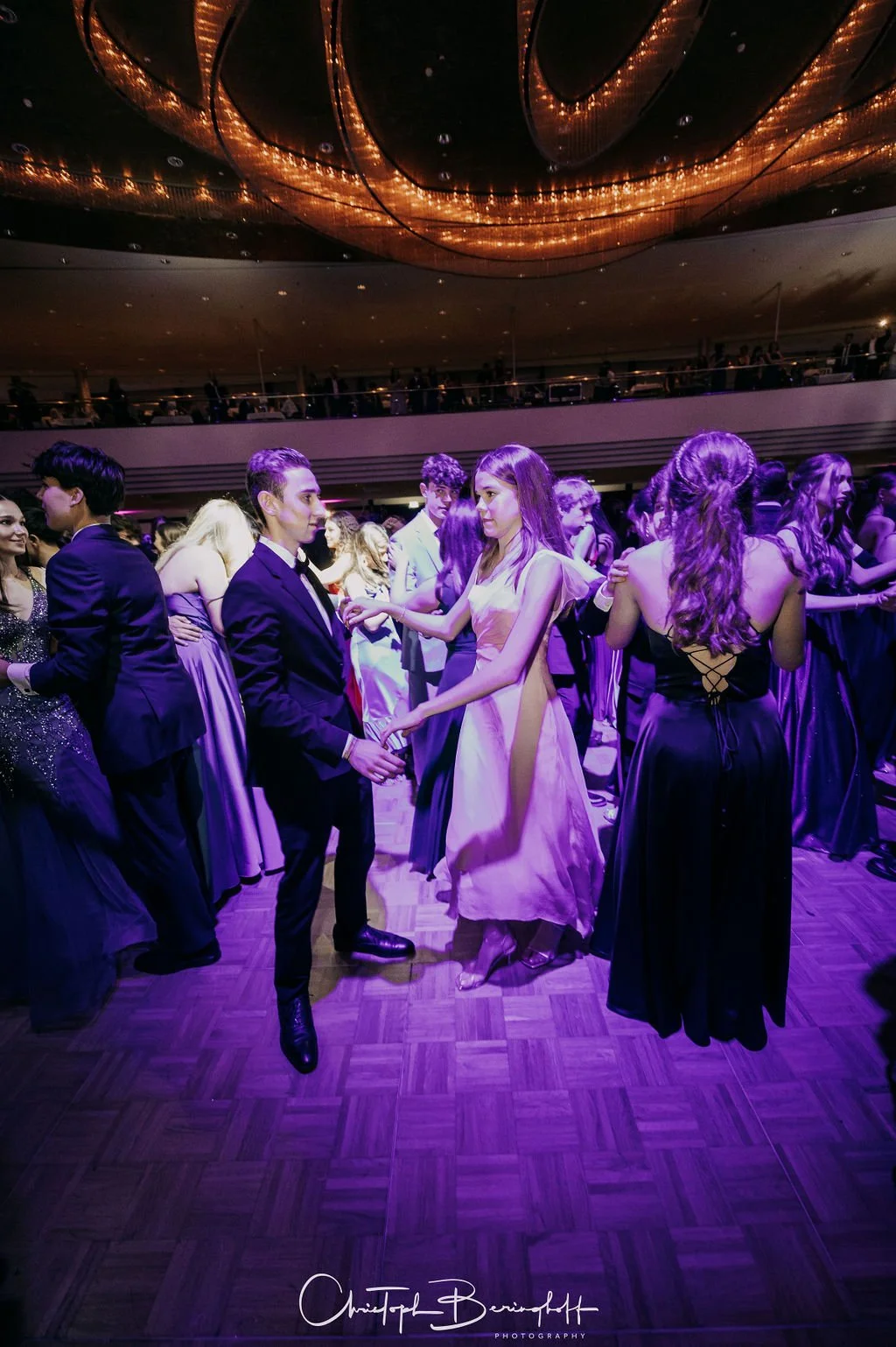 Junge Menschen tanzen auf einem Ball in formeller Abendkleidung in einem Veranstaltungsraum mit großen, warm beleuchteten Decken. Die Stimmung ist festlich und elegant.