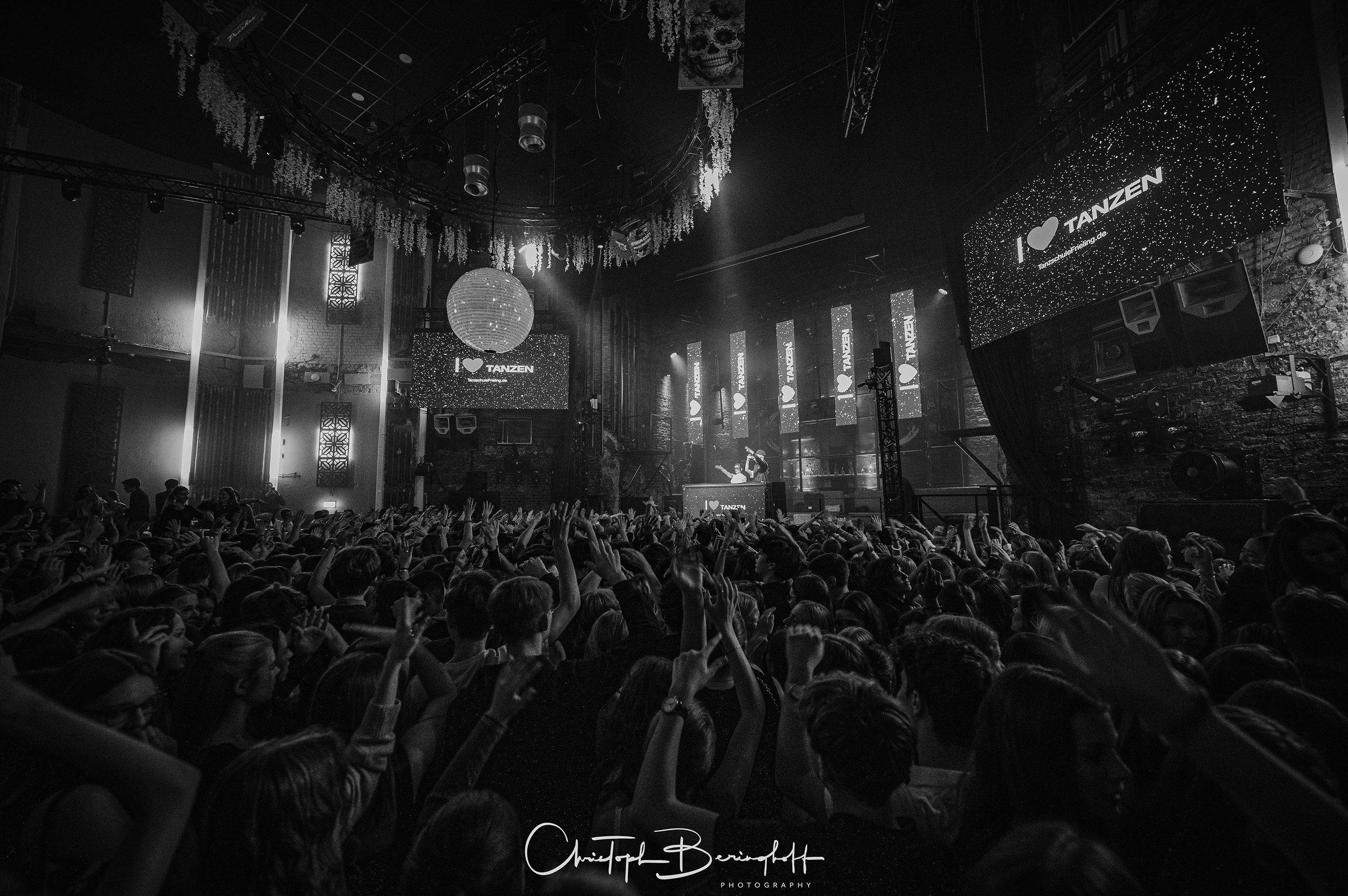 Ein thousands of Menschen tanzen und feiern in einem Konzertsaal mit mehreren Bildschirmen, einer großen hängenden Laterne und beeindruckender Beleuchtung.