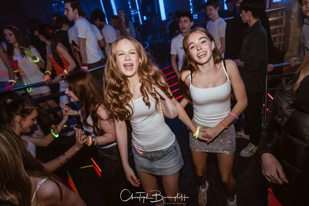 Two smiling young girls are dancing in a disco lit by neon lights, surrounded by other teenagers on the dance floor.