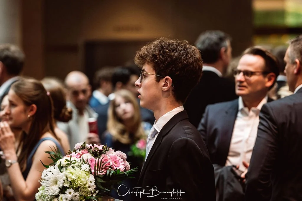Eine junge Frau mit Kurzhaarfrisur, Brille und Anzug hält einen Blumenstrauß bei einer Veranstaltung. Im Hintergrund sind andere Menschen in formeller Kleidung zu sehen.