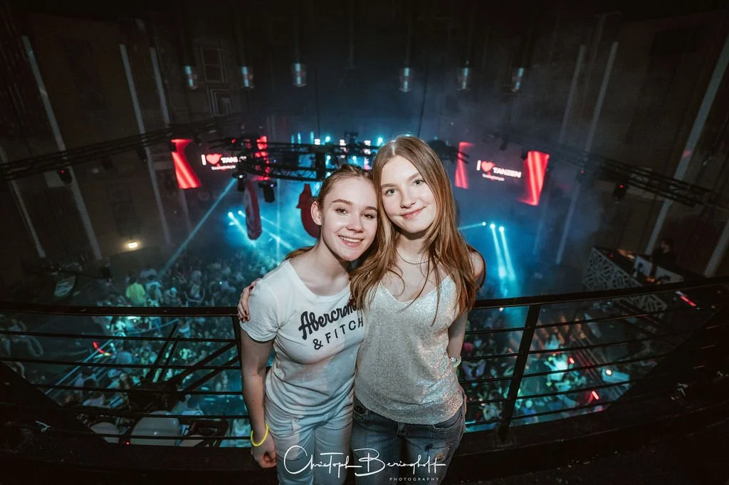 Two young women are standing on a stage or terrace in front of a party with strobe lights and laser effects; in the background is a dance floor full of people.