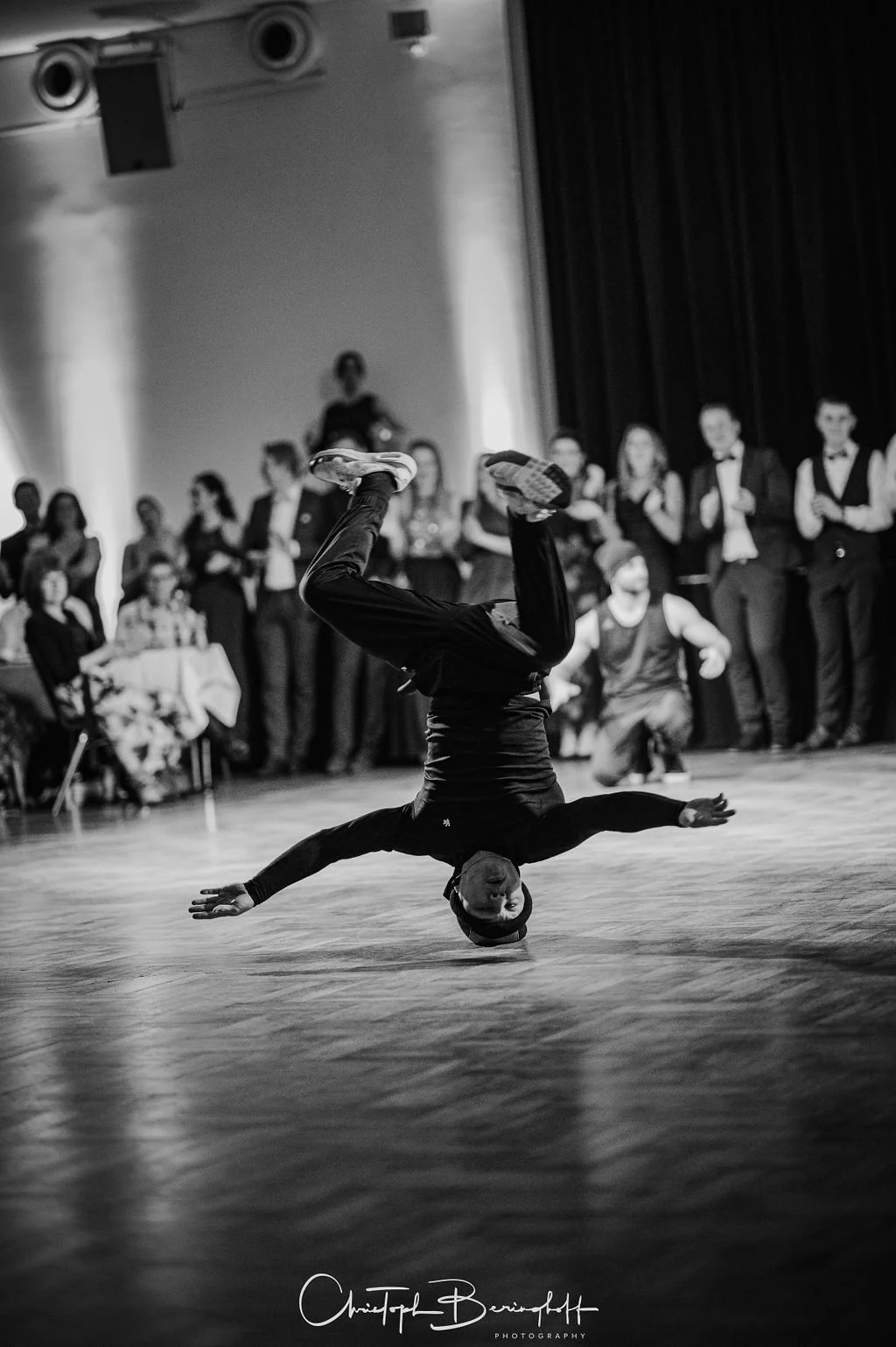 Breakdancer performance bei einem Wettbewerb im schwarzen Hemd und schwarzen Hosen, kämpft mit einem Handstand auf dem Boden, während eine Gruppe von Gästen in formeller Kleidung im Hintergrund zuschaut.