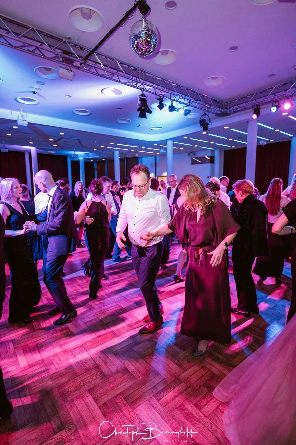 Menschen tanzen auf einer Party in einem festlich dekorierten Raum mit Diskokugel an der Decke und bunter Beleuchtung.