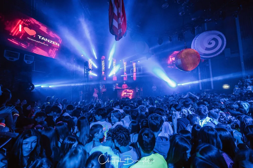 A crowd at a dance party with colorful lighting and disco balls.