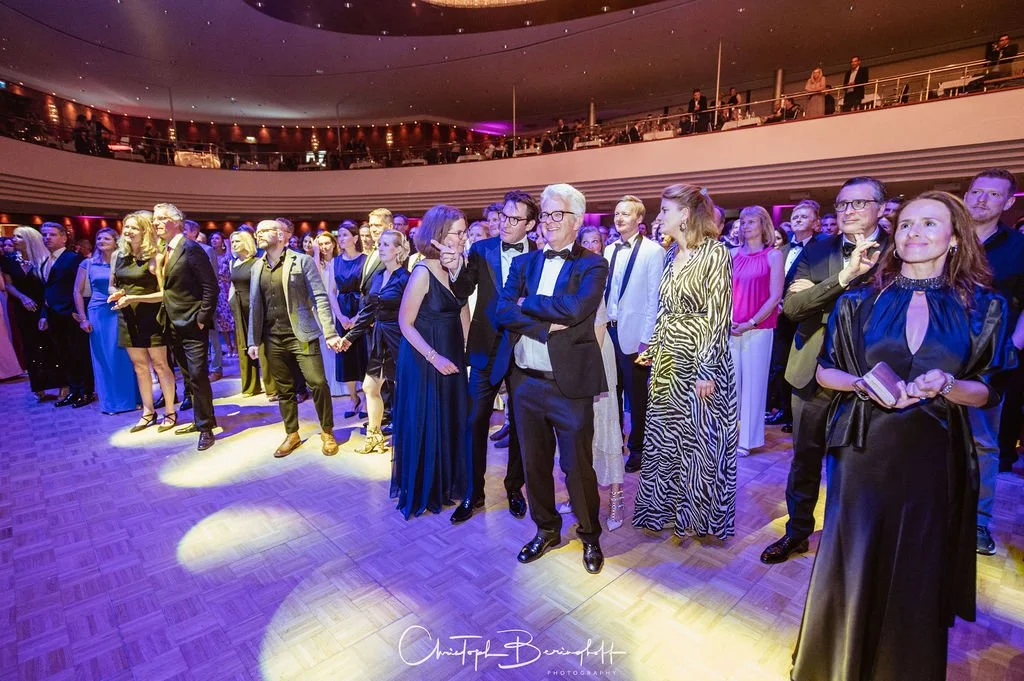 Menschen in eleganter Abendgarderobe auf einer Ballsaal-Bühne bei einer formellen Veranstaltung, mit einigen Gesprächen und Blicken in verschiedene Richtungen.