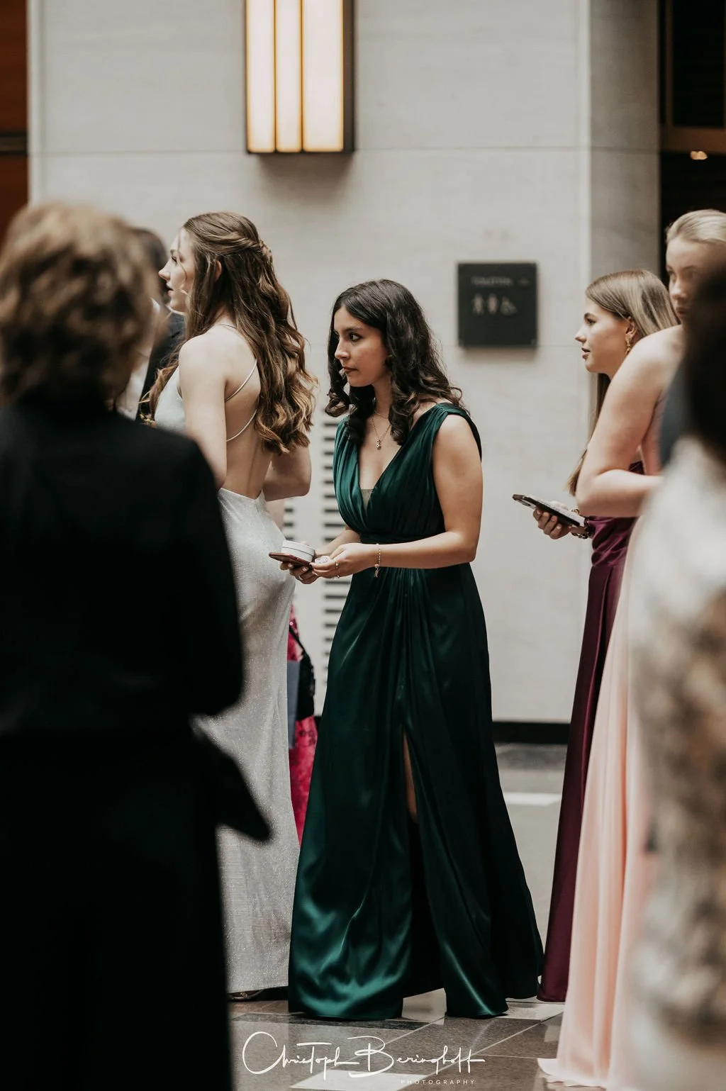 Gruppe von Frauen in eleganten Kleidern bei einer formellen Veranstaltung, die in einem modernen Gebäude mit beleuchteter Wandinstallation stehen.