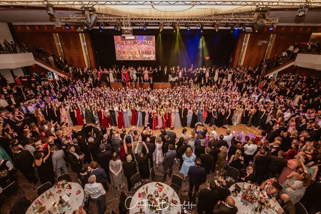 Festliche Ballnacht in einem großen Saal mit vielen Gästen, romantischer Dekoration, Stühlen und Tischen, Tanzfläche, Bühnenbild, und einer großen Gruppe von Menschen in eleganten Kleidern und Anzügen.
