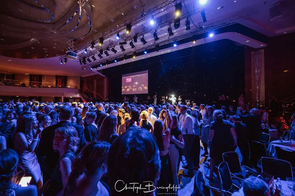 Menschen, die auf einer Tanzfläche in einem eleganten Veranstaltungsraum tanzen, mit Bühne und Leinwand im Hintergrund, in festlicher Atmosphäre bei einer Gala oder einem Ball.
