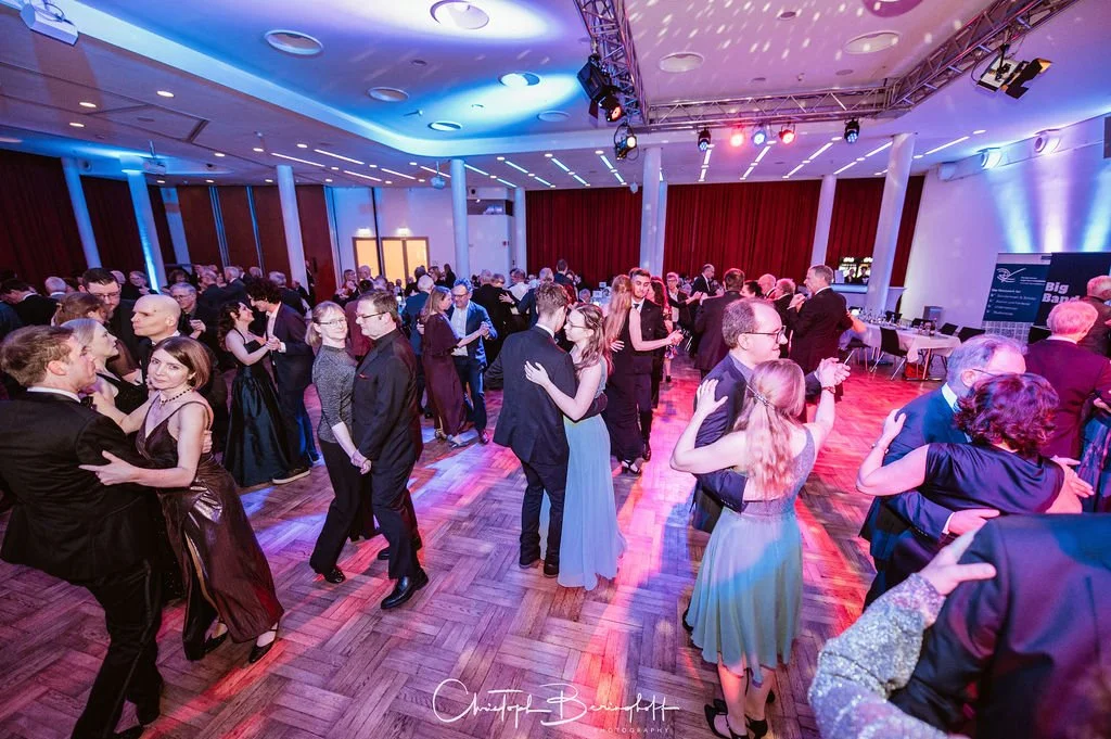 Viele Menschen tanzen elegant auf einer festlichen Veranstaltung in einem großen Saal mit Holzparkett, buntem Deckenlicht und einem Bühnenbereich.
