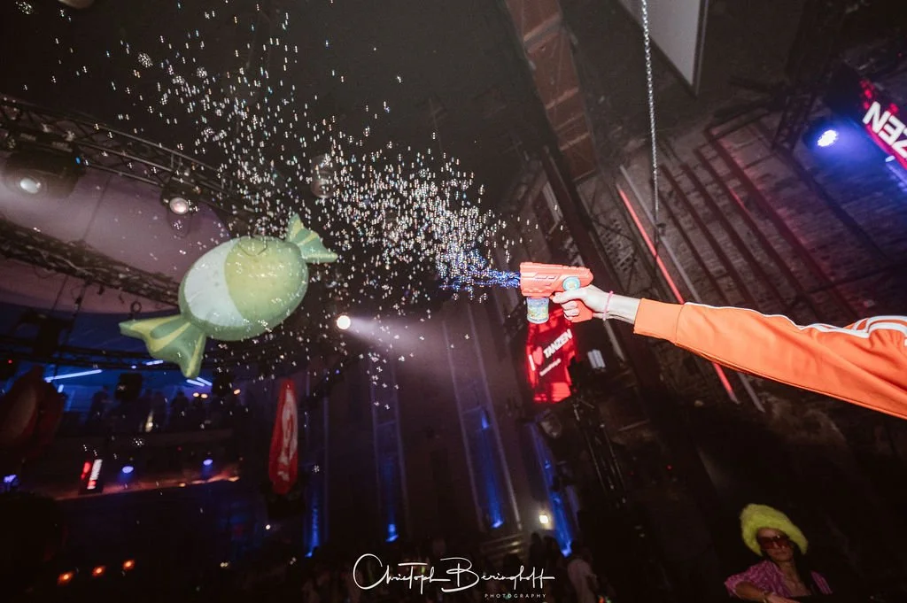 A person is spraying a toy gun at a decorative light fixture hanging from the ceiling that looks like a fish, in a nightclub or party venue with dim lighting and several people in the background.